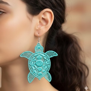 Może przedstawiać: Turkusowy, ażurowy kolczyk w kształcie żółwia. Kolczyk wykonany jest z delikatnego, haftowanego materiału. Wzór żółwia ma szczegółowy pancerz i płetwy, ze srebrnym haczykiem.