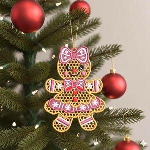 Peut inclure: Ornement de Noël en forme de bonhomme en pain d'épices doré, avec un nœud et une robe roses, et un visage souriant. L'ornement est suspendu à un sapin de Noël avec des boules rouges.