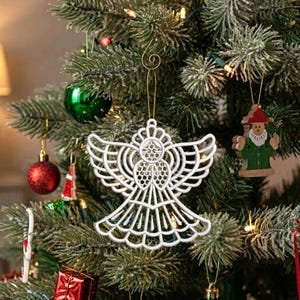 May include: A white, openwork angel ornament with intricate, symmetrical geometric patterns and a circular halo, suspended by a gold string. The angel features a wide, flared skirt and outstretched wings, hanging from a green pine tree branch.