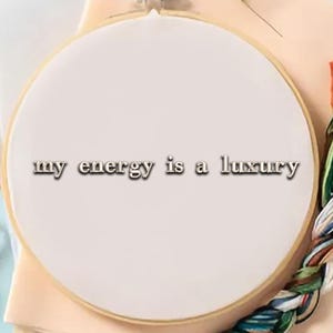 May include: A white embroidery hoop with the text "my energy is a luxury" in a serif font. The hoop is set on a beige fabric, with colorful embroidery floss and pink scissors visible.