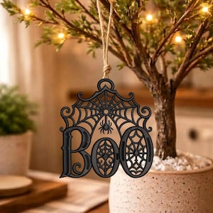 May include: A black laser-cut wooden ornament featuring the word BOO in a decorative font, topped with an intricate spiderweb design and a small spider hanging from the center, suspended by a thin tan twine string for hanging display.