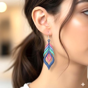 May include: A close-up of a colorful feather-shaped earring. The earring features a layered design with shades of blue, pink, and green. The earring is suspended from a silver hook. The earring is a unique piece of jewelry.