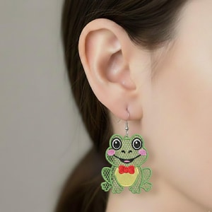 May include: A pair of whimsical frog earrings. Each earring features a green frog design with a yellow belly, a red bow tie, and pink cheeks. The earrings are suspended from silver-colored hooks.