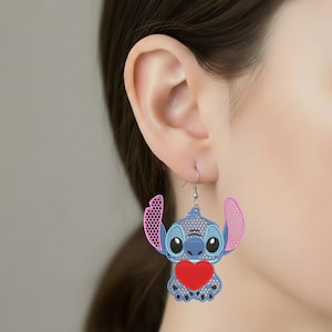 Peut inclure: Boucles d'oreilles Stitch bleues et roses avec un cœur rouge. Les boucles d'oreilles présentent un motif de personnage de dessin animé avec un motif ajouré détaillé. Les boucles d'oreilles sont suspendues à un crochet argenté.
