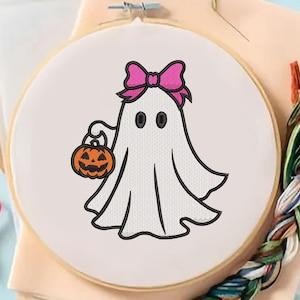 May include: An embroidery hoop featuring a white ghost with a pink bow, holding an orange jack-o'-lantern. The ghost has black eyes and a black outline. The design is set against a white fabric background, with pink scissors and thread spools visible.