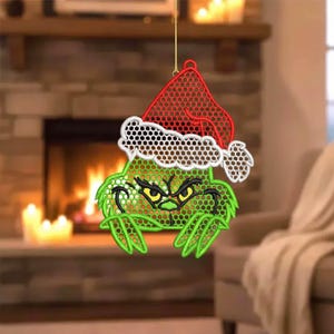 May include: A green Christmas ornament featuring a cartoon character with a red Santa hat and a white trim. The ornament has a mesh-like design and is hanging in front of a fireplace.