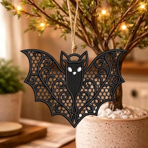 May include: A black bat-shaped ornament featuring intricate lace-like embroidery with a honeycomb pattern and swirling lines. The bat has two small white eyes and is suspended by a thin, light brown jute twine loop attached to the top center.