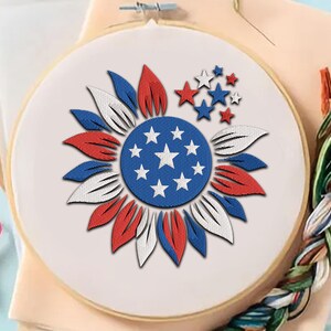 Könnte beinhalten: Gesticktes Sonnenblumen-Design in einem Holzreifen. Die Sonnenblume hat rote, weiße und blaue Blütenblätter und eine blaue Mitte mit weißen Sternen. Zusätzliche rote und blaue Sterne befinden sich über der Blume. Bastelbedarf ist im Hintergrund sichtbar.