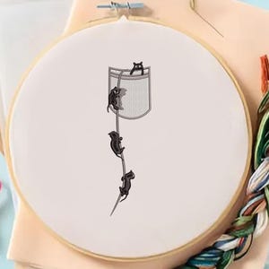May include: Embroidery hoop featuring a whimsical design of black cats climbing a rope towards a pocket. The cats are embroidered on a white fabric, set within a wooden hoop. Craft supplies, including thread and scissors, are visible.