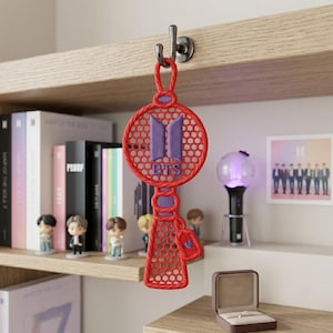 May include: A red and purple embroidered BTS logo keychain hanging from a metal hook. The design features a circular top with a mesh pattern, a central purple BTS logo, and a rectangular base with a matching mesh texture and a small heart charm.
