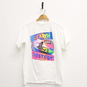 May include: White t-shirt featuring a colorful graphic of a surfer riding a wave. The design includes the words "SEARCH AND DESTROY" in a vibrant, retro style. The surfer is depicted in motion on a green surfboard.