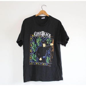 Peut inclure: Un t-shirt noir à manches courtes avec une impression graphique de Clint Black portant un chapeau de cowboy. Le motif comprend des cactus verts, un fond bleu tourbillonnant et une bordure décorative avec de petites formes jaunes et bleues.
