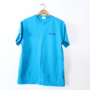 May include: A turquoise blue t-shirt with the word "Microsoft" printed in black on the upper chest. The short-sleeved shirt is hanging on a wooden hanger against a white background.