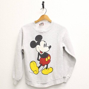 Peut inclure: Un sweat-shirt gris clair avec un grand dessin de Mickey Mouse. Mickey est représenté dans sa pose classique, portant un short rouge, des chaussures jaunes et des gants noirs. Le sweat-shirt a un col rond et des manches longues.
