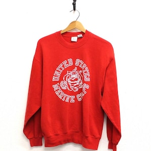 May include: A red sweatshirt with a white graphic of a bulldog and the words "United States Marine Corps" in a circular design. The sweatshirt has long sleeves and a crew neck.