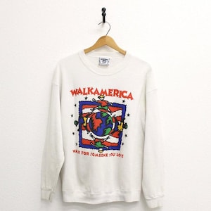 May include: A white long-sleeve crewneck sweatshirt featuring a graphic print with the text WALKAMERICA and WALK FOR SOMEONE YOU LOVE. The design includes a central globe surrounded by stylised figures, a blue and red border, and black stars.