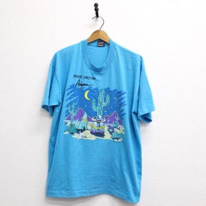 May include: A turquoise t-shirt featuring a desert landscape design with a large cactus, a crescent moon, and the text "Apache Junction Arizona." The graphic is in shades of blue, purple, and yellow.