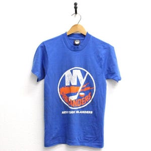 May include: A royal blue short-sleeve t-shirt featuring a circular white logo on the chest. The logo contains the letters NY in white, an orange hockey stick, and the word ISLANDERS in orange. The text NEW YORK ISLANDERS appears below the logo.
