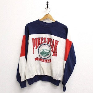 Op de afbeelding: Een vintage crewneck sweatshirt met een colorblock design. Het bovenste gedeelte is marineblauw, het midden is wit met een "Pikes Peak Colorado" graphic, en de mouwen hebben rode accenten. De sweatshirt hangt aan een houten hanger.