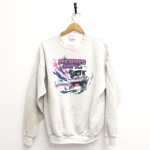 May include: A light gray sweatshirt with a crew neck and long sleeves. The front features a graphic of a vehicle with the text "DENNIS KIRK" in purple and pink, with the word "Performance" below. The design includes splashes of pink and blue.