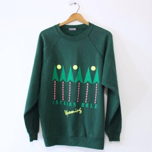 Op de afbeelding: Een donkergroene sweatshirt met een grafisch ontwerp van groene bomen, gele cirkels en roze verticale lijnen. De tekst "JACKSON HOLE Wyoming" staat eronder in het geel gedrukt. De sweatshirt hangt aan een houten hanger.