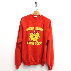 May include: A red sweatshirt with the words "UNITED STATES MARINE CORPS" in yellow lettering. A yellow bulldog wearing a hat is in the center. The sweatshirt is hanging on a wooden hanger.