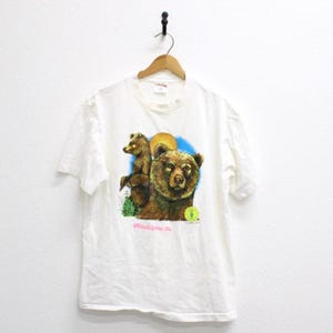 Peut inclure: T-shirt blanc avec un motif de deux ours bruns, un soleil jaune et un ciel bleu. Le texte "Philadelphia, Pa" est imprimé sous les ours. Le t-shirt a des manches courtes et un col rond.