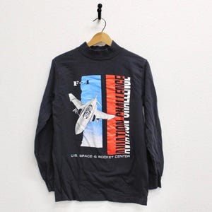 May include: Black long-sleeve shirt with a mock neck. The shirt features a graphic of a jet plane with the text "F-4" and "AVIATION CHALLENGE". The text "U.S. SPACE & ROCKET CENTER" is also printed on the shirt.