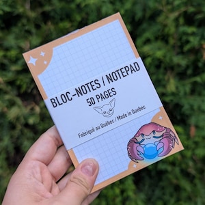 Peut inclure: Un bloc-notes avec un motif quadrillé et le texte "BLOC-NOTES / NOTEPAD 50 PAGES". La couverture présente un crabe de dessin animé tenant un globe bleu brillant. Le bloc-notes est présenté sur un fond de feuillage vert.