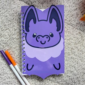 May include: A spiral-bound notebook with a purple bat illustration on the cover. The bat has large ears and a smiling face. The notebook is next to three colorful markers: purple, orange, and white.