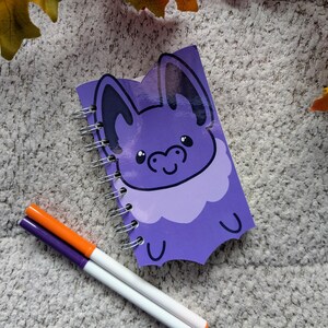 Peut inclure: Un carnet violet à spirales avec un dessin de chauve-souris de dessin animé. La chauve-souris a de grandes oreilles et un visage souriant. Deux marqueurs, un orange et un violet, sont placés près du carnet. Le fond est une surface texturée gris clair.
