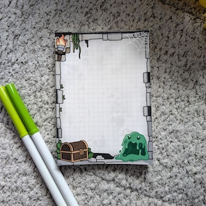 May include: A rectangular notepad with a dungeon-themed border, featuring a treasure chest, green slime, and a torch. Two green and white markers are positioned near the notepad. The notepad has a grid pattern.