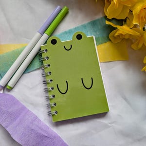 May include: A green frog-faced notebook with a spiral binding, two pens, and colourful crepe paper on a white surface. Yellow flowers are in the background. The notebook features a smiling face and two curved lines.