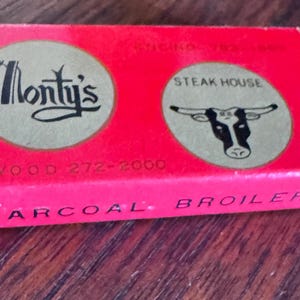 May include: A rectangular red matchbox featuring two circular gold logos. The left logo displays the text Monty's in black script. The right logo shows a black and white bull head with the text STEAK HOUSE. Text reads WESTWOOD 272-2000 and CHARCOAL BROILER.