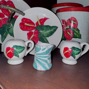May include: A collection of kitchenware featuring white ceramic plates, a canister with a red lid, and two round salt and pepper shakers, all decorated with red anthurium flowers and green leaves, alongside a small creamer with a teal leaf pattern.