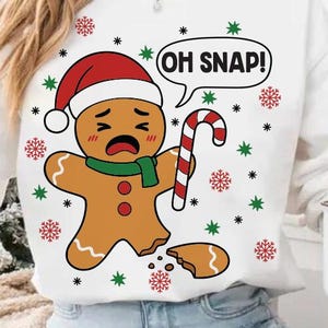 May include: White sweatshirt featuring a distressed gingerbread man wearing a Santa hat and green scarf, holding a candy cane. The text "OH SNAP!" is in a speech bubble. Red and green snowflake and star accents.