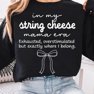 May include: Black sweatshirt with white text. The text reads "in my string cheese mama era" with the phrase "Exhausted, overstimulated but exactly where I belong" below. A white bow graphic is below the text.