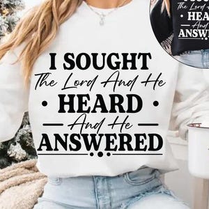 Puede incluir: Una sudadera blanca con el texto "I SOUGHT The Lord And He HEARD And He ANSWERED" en negro. La imagen también muestra una taza con un diseño de Papá Noel y un árbol de Navidad en el fondo.