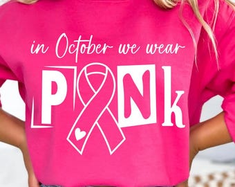 May include: A bright pink sweatshirt with the text "in October we wear PINK" in white. The word "PINK" is stylized with a white ribbon and a heart. The sweatshirt is paired with blue denim jeans.