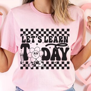 Op de afbeelding: Lichtroze t-shirt met de zwarte tekst "LET'S LEARN TODAY", samen met een cartoon bloem en potlood afbeelding. Het ontwerp bevat een zwart-wit geblokt patroon boven en onder de tekst.