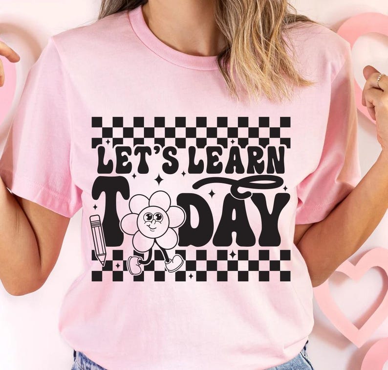 Op de afbeelding: Lichtroze t-shirt met de zwarte tekst "LET'S LEARN TODAY", samen met een cartoon bloem en potlood afbeelding. Het ontwerp bevat een zwart-wit geblokt patroon boven en onder de tekst.
