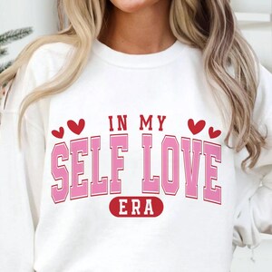May include: A white sweatshirt with the text "IN MY SELF LOVE ERA" in pink block letters with a red outline. Two red hearts are above the words "IN MY" and below the word "ERA".