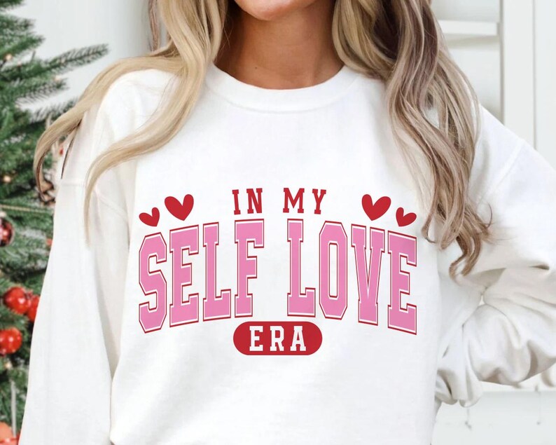 May include: A white sweatshirt with the text "IN MY SELF LOVE ERA" in pink block letters with a red outline. Two red hearts are above the words "IN MY" and below the word "ERA".
