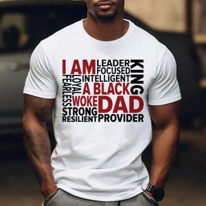 Op de afbeelding: Wit T-shirt met de tekst "I AM A BLACK WOKE DAD" in een gelaagd ontwerp. Het shirt toont ook de woorden "Leader, Focused, Intelligent, Fearless, Loyal, Strong, Resilient, Provider, King".