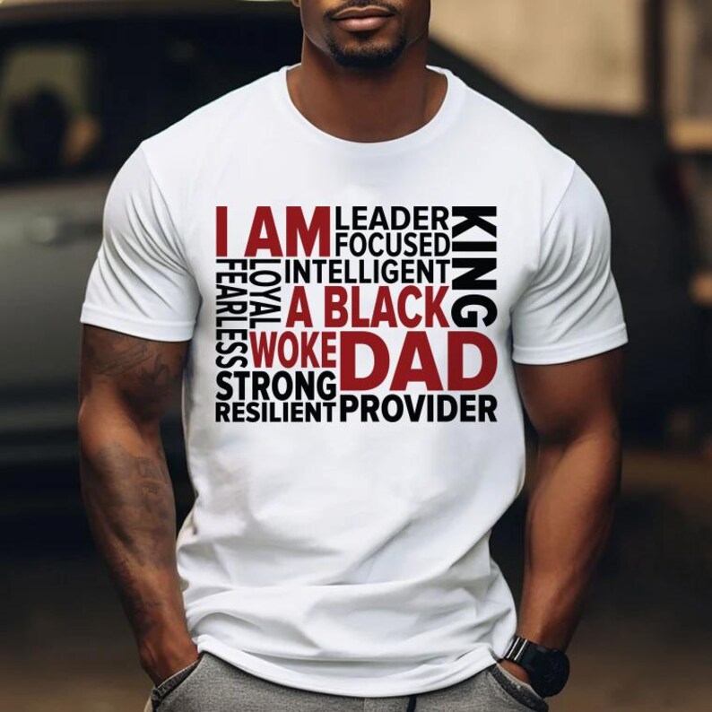 Op de afbeelding: Wit T-shirt met de tekst "I AM A BLACK WOKE DAD" in een gelaagd ontwerp. Het shirt toont ook de woorden "Leader, Focused, Intelligent, Fearless, Loyal, Strong, Resilient, Provider, King".