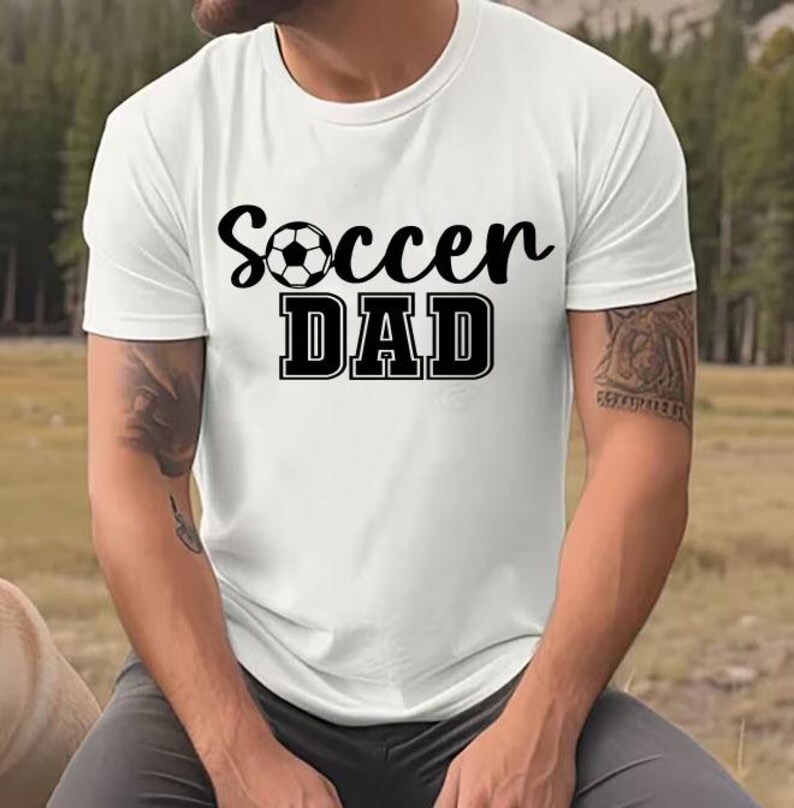 May include: White t-shirt with black text that reads "Soccer Dad" and a black and white soccer ball graphic.
