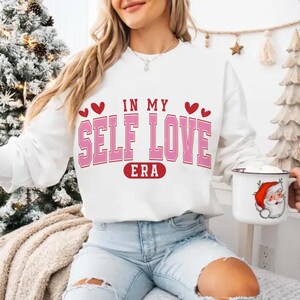 May include: White sweatshirt with the text "IN MY SELF LOVE ERA" in pink and red. The sweatshirt has a crew neck and long sleeves. The model is wearing light blue jeans and holding a mug with a Santa Claus design.