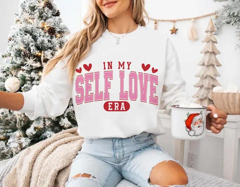 May include: White sweatshirt with the text "IN MY SELF LOVE ERA" in pink and red. The sweatshirt has a crew neck and long sleeves. The model is wearing light blue jeans and holding a mug with a Santa Claus design.