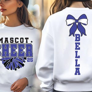 May include: White sweatshirt with a blue bow and the word "BELLA" spelled vertically down the back. The front of the sweatshirt has a black and white cheerleading pom-pom graphic and the text "MASCOT CHEER 20 25".