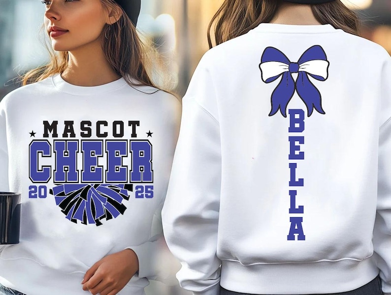 May include: White sweatshirt with a blue bow and the word "BELLA" spelled vertically down the back. The front of the sweatshirt has a black and white cheerleading pom-pom graphic and the text "MASCOT CHEER 20 25".